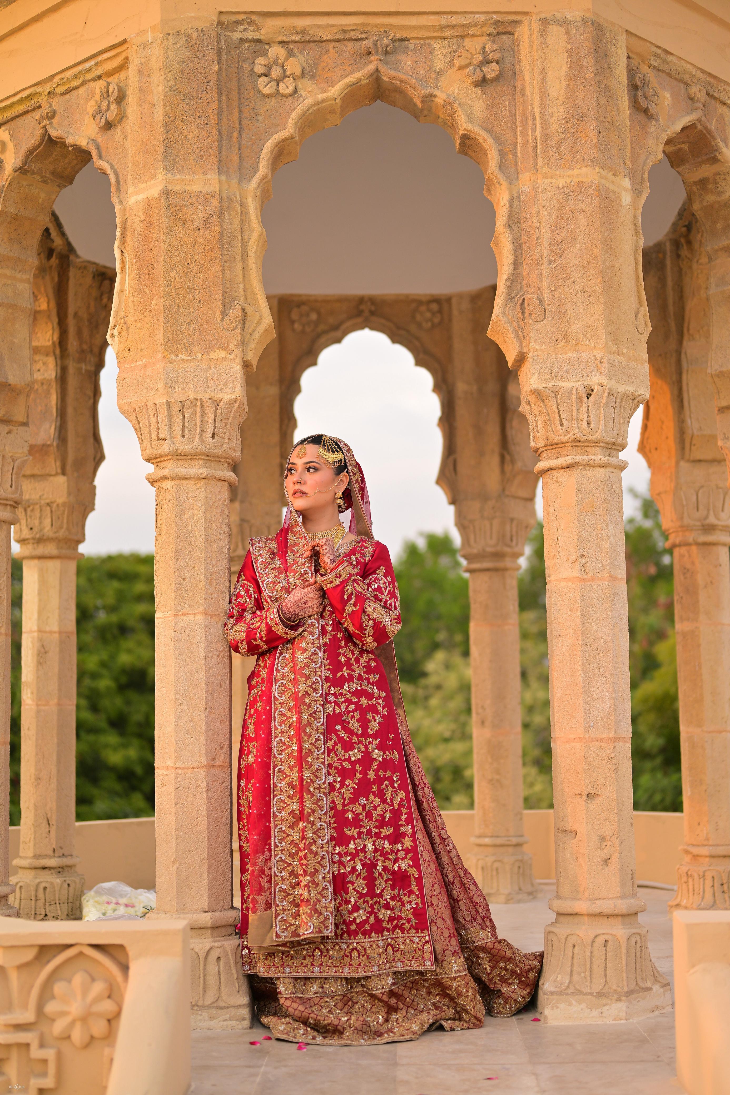 Real Sameera's Collection bride in deep red and gold bridal couture beneath Mughal sandstone arches at golden hour